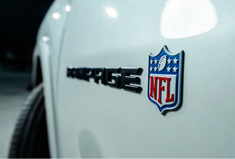 Foco na lateral dianteira de picape branca Ram Rampage R/T NFL Edition mostrando símbolo da NFL, que aparece nas cores originais, branco, azul e vermelho, em alto relevo. O símbolo possui formato de escudo, quatro estrelas, uma bola de futebol americano e as letras NFL. Ao lado, também em alto relevo, porém desfocado, está escrito Rampage, em preto. 