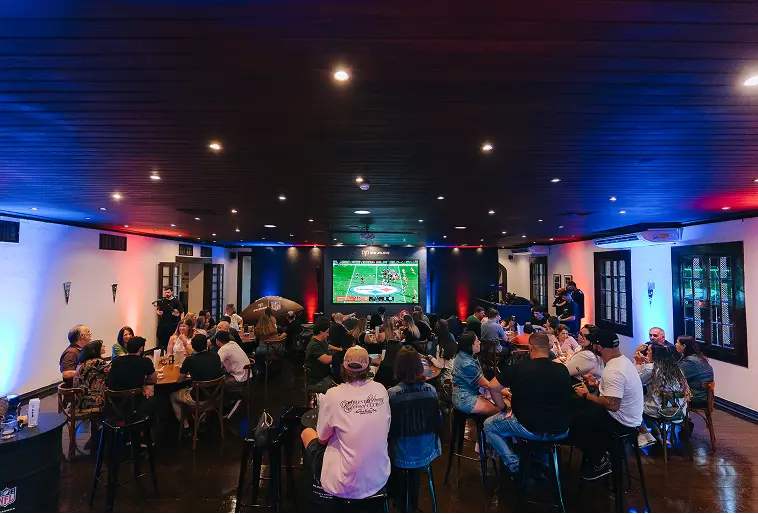 Ambiente escuro com luzes próximas ao teto nas cores azul e vermelho. Diversas pessoas estão no ambiente, que tem tom festivo e possui enorme telão com a transmissão de um jogo de futebol americano.