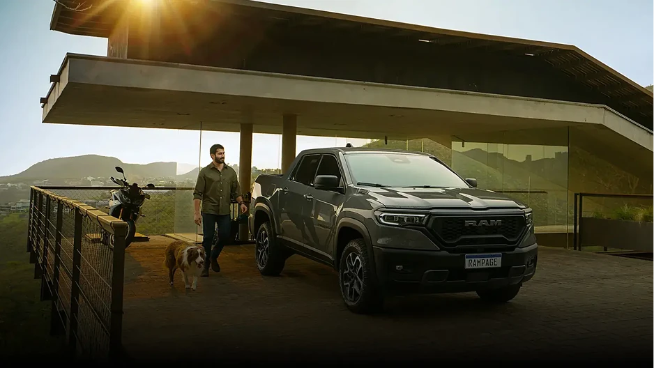 Foto de vista lateral-frontal de picape Ram de cor cinza estacionada em garagem de luxo ao lado de homem segurando um cão São Bernado pela guia ao lado esquerdo.
